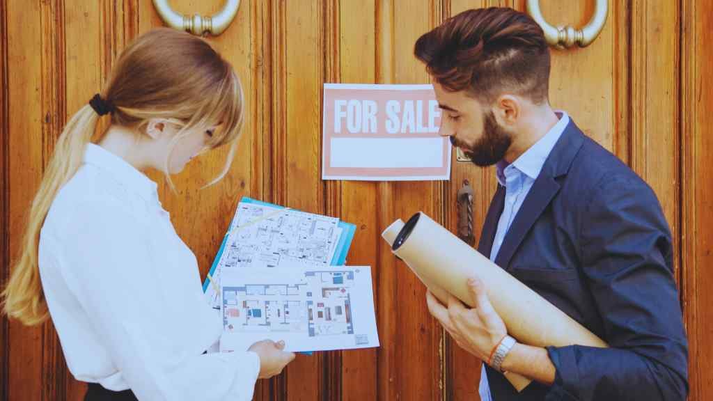 A man and woman stand in front of a wooden door with a "For Sale" sign, examining blueprints. The scene conveys a professional, focused atmosphere.