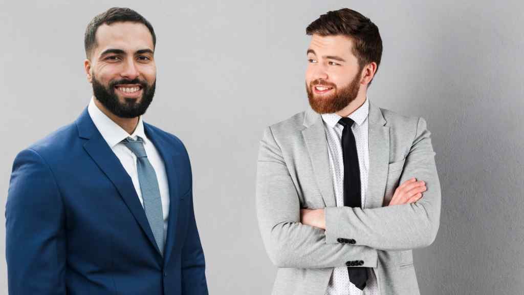 Two men in business attire stand against a gray background. The man on the left wears a blue suit and smiles confidently. The man on the right, in a gray suit, looks at him with a grin.