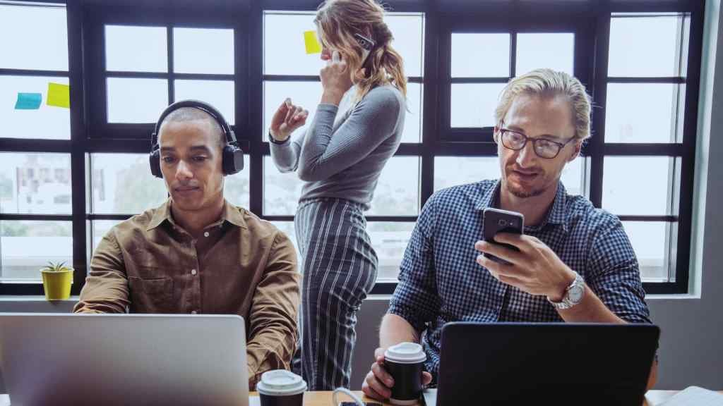 Three professionals work at desks with laptops. One wears headphones, another looks at a phone, and a woman is on a call. The scene is busy yet focused.