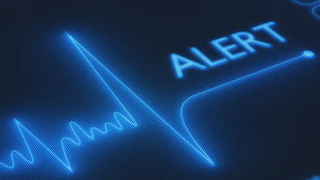 Glowing blue electrocardiogram line with a jagged peak on a dark background, next to the word "ALERT" in capital letters. Conveys urgency.