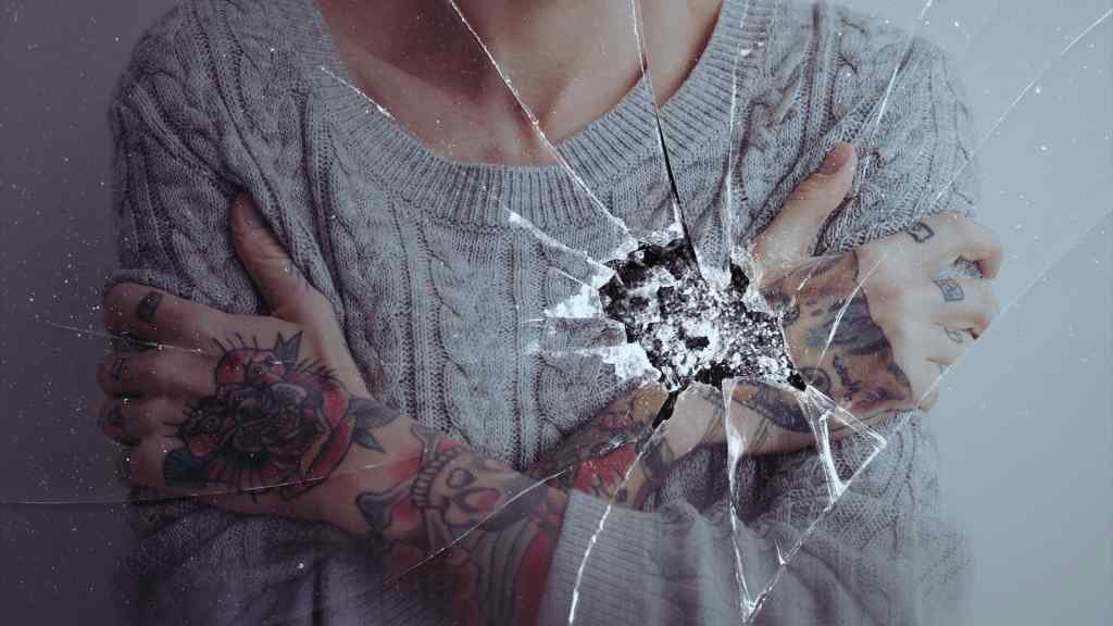 A person in a gray cable-knit sweater, arms crossed, displays colorful tattoos. The image features a cracked glass effect, conveying a sense of vulnerability.