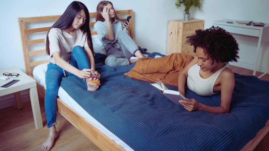 Three young women relax in a cozy bedroom. One reads a book, another checks her phone, and the third adjusts her sock, creating a calm, casual vibe.